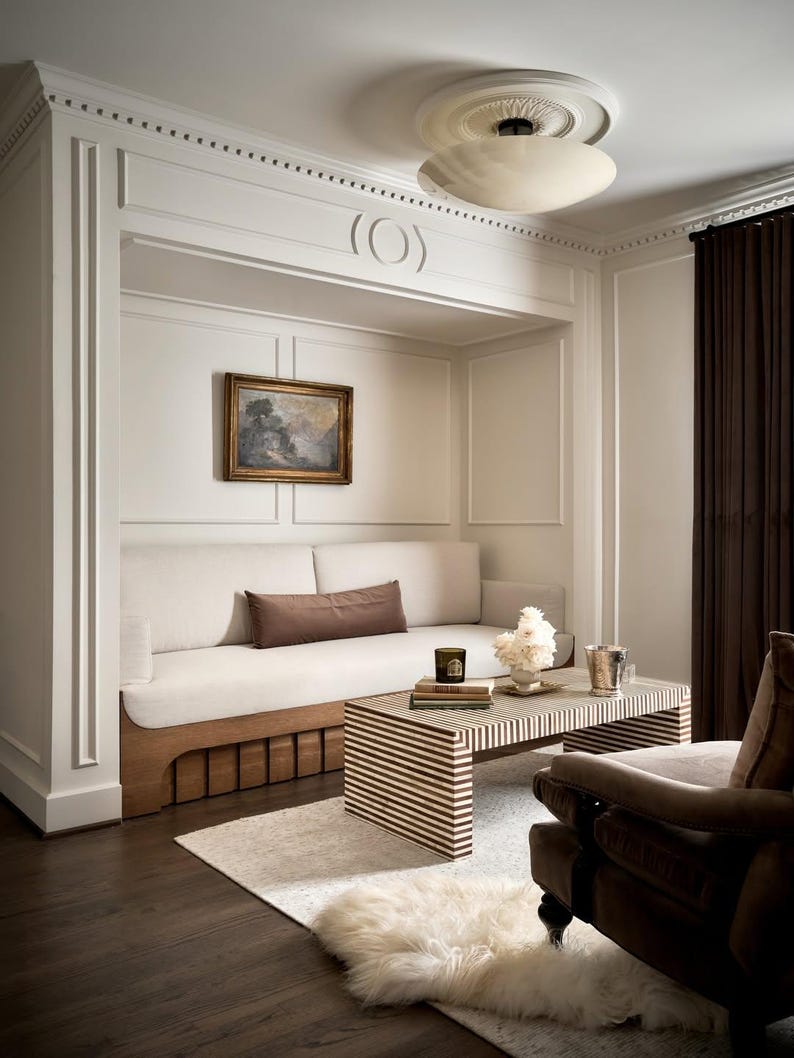 K&ouml;nnte beinhalten: Ein gem&uuml;tliches Wohnzimmer mit einem eingebauten wei&szlig;en Sofa, einem gestreiften Couchtisch und einem braunen Sessel. Der Raum verf&uuml;gt &uuml;ber ein kleines Gem&auml;lde, eine Deckenleuchte und dunkelbraune Vorh&auml;nge. Ein flauschiger wei&szlig;er Teppich liegt auf dem Boden.