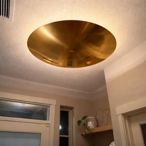 K&ouml;nnte beinhalten: Eine gro&szlig;e, kreisf&ouml;rmige Deckenleuchte in Goldfarbe. Das Licht ist in die Decke eingelassen und hat eine glatte, reflektierende Oberfl&auml;che. Die Leuchte ist der Mittelpunkt des Raumes und strahlt ein warmes Licht aus.