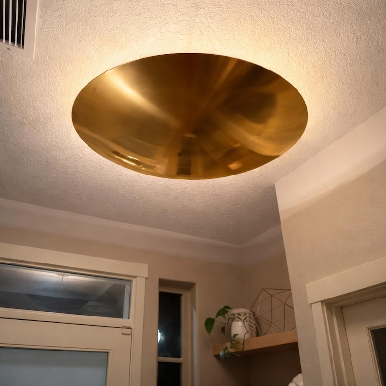 K&ouml;nnte beinhalten: Eine gro&szlig;e, kreisf&ouml;rmige Deckenleuchte in Goldfarbe. Das Licht ist in die Decke eingelassen und hat eine glatte, reflektierende Oberfl&auml;che. Die Leuchte ist der Mittelpunkt des Raumes und strahlt ein warmes Licht aus.