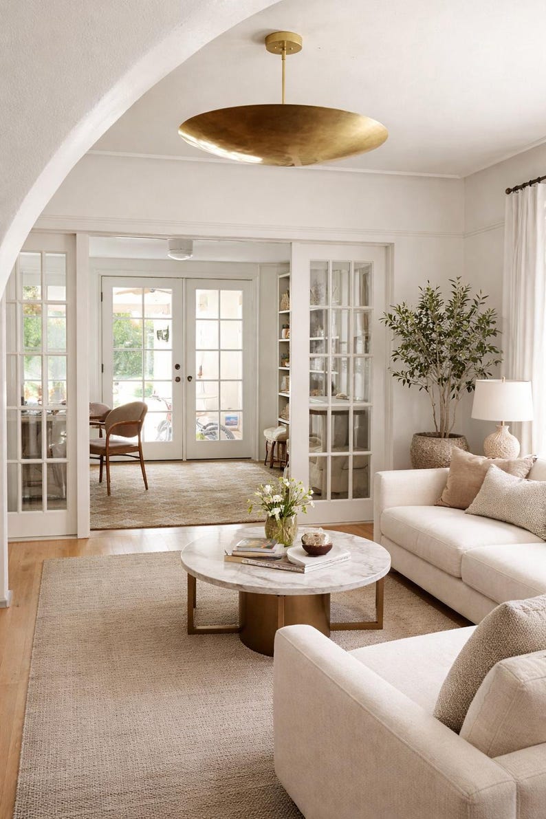 K&ouml;nnte beinhalten: Helles, luftiges Wohnzimmer mit neutraler Farbpalette. Es verf&uuml;gt &uuml;ber ein wei&szlig;es Sofa mit Zierkissen, einen Couchtisch mit Marmorplatte und eine gro&szlig;e goldene Pendelleuchte. Eine Topfpflanze und ein Teppich sorgen f&uuml;r Textur.