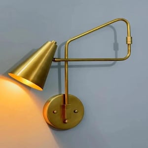 May include: A gold-colored wall sconce with a conical shade and an adjustable arm. The sconce has a circular base and a warm light emanating from the shade. The design is modern and minimalist.