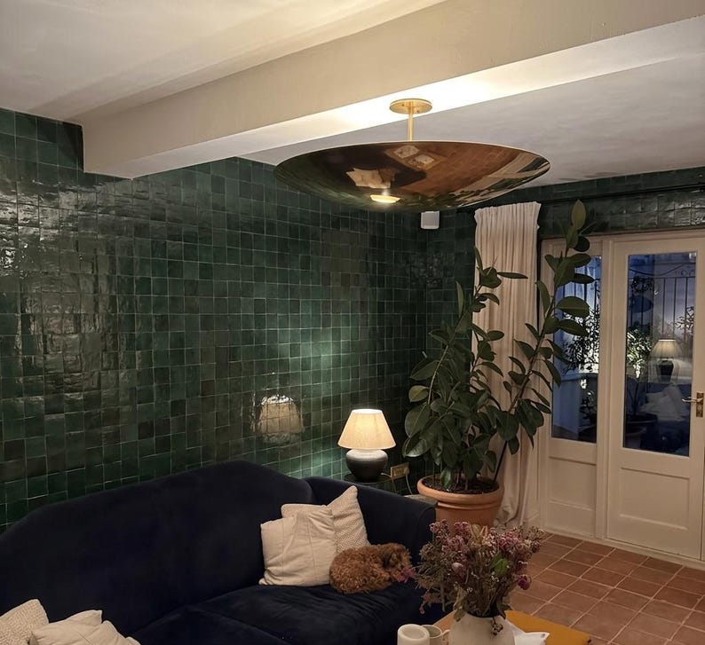 K&ouml;nnte beinhalten: Ein modernes Wohnzimmer mit einem dunkelblauen Sofa, wei&szlig;en Kissen und einer gro&szlig;en, reflektierenden goldfarbenen Deckenleuchte. Die W&auml;nde sind mit dunkelgr&uuml;nen Fliesen bedeckt. Eine Topfpflanze und ein kleiner Hund sind ebenfalls zu sehen.