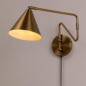 May include: A brass wall sconce featuring a conical lampshade and an adjustable arm. The lamp has a circular base and a fabric-covered cord. The light is on, illuminating the interior of the shade.