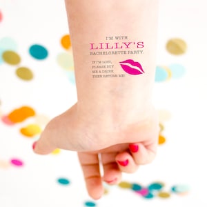 Può includere: Un tatuaggio temporaneo su un polso con la scritta "I'M WITH LILLY'S BACHELORETTE PARTY." Il tatuaggio include anche la scritta "IF I'M LOST, PLEASE BUY ME A DRINK THEN RETURN ME!" e una grafica di labbra rosa. Le unghie della persona sono dipinte di rosso.