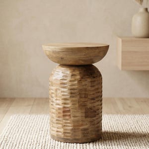 May include: A light brown wooden side table with a carved, textured base and a round top. The table is approximately 46 cm tall and sits on a woven rug. The background includes a vase with dried plants.