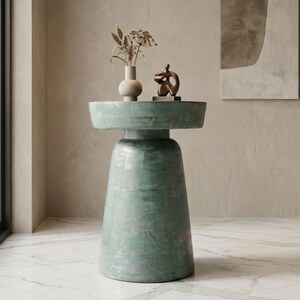 May include: A pale green side table with a circular top and a pedestal base. It features a small vase with dried flowers and a bronze sculpture. The table is set on a marble floor.