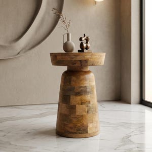 May include: A wooden side table with a round top and a wide, tapered base. The table is made of light brown wood with a patchwork design. A small vase with a sprig of greenery and a small sculpture sit on top of the table.