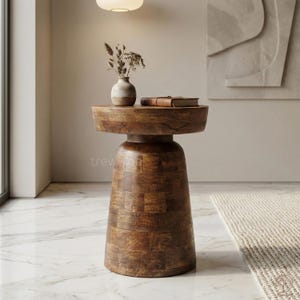 Puede incluir: Una mesa auxiliar redonda de madera con base de pedestal. La mesa es de madera marrón oscuro y tiene un jarrón decorativo con flores secas y un libro encima. La mesa mide aproximadamente 61 cm de altura.