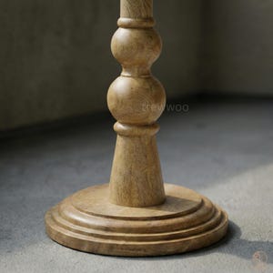 May include: A light brown wooden table base with a round, tiered foot and a turned, decorative column. The column has a bulbous design with a smooth, natural finish. The text "trewwoo" is visible on the column.