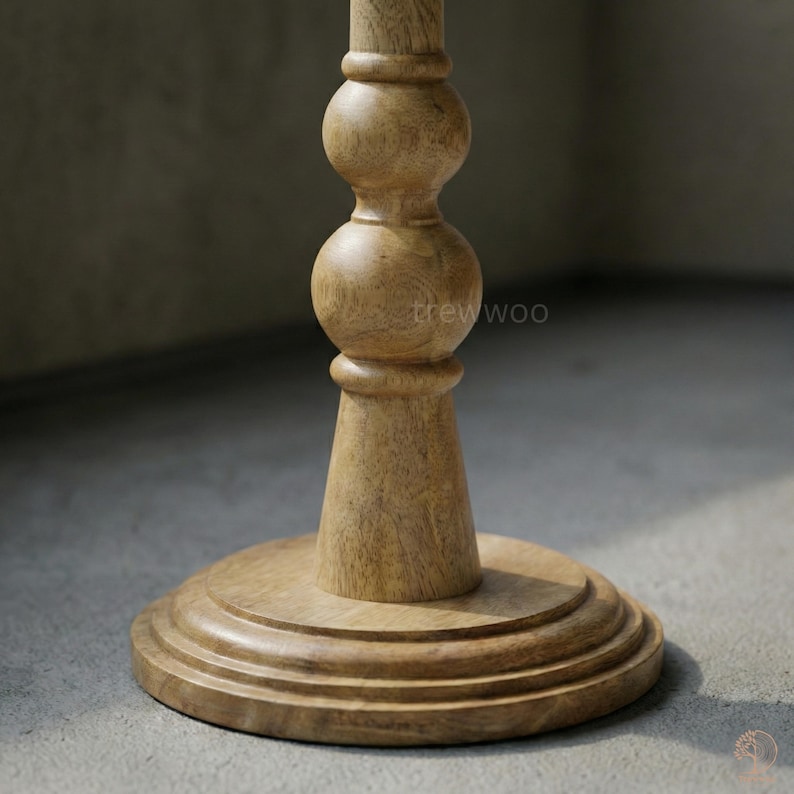May include: A light brown wooden table base with a round, tiered foot and a turned, decorative column. The column has a bulbous design with a smooth, natural finish. The text "trewwoo" is visible on the column.