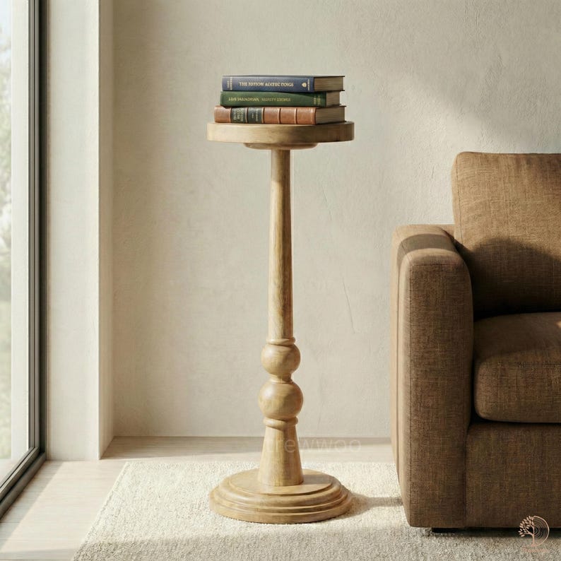 May include: A tall, light-colored wooden side table with a round top and base. The table has a slender, turned column with two decorative spheres. Three books are stacked on the top. The table is next to a brown sofa.