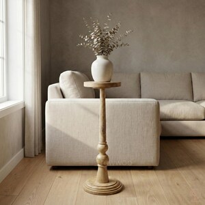 May include: A tall, light-colored wooden side table with a round top and base. A white ceramic vase with dried foliage sits on the table. The table is in front of a beige sofa in a well-lit room.