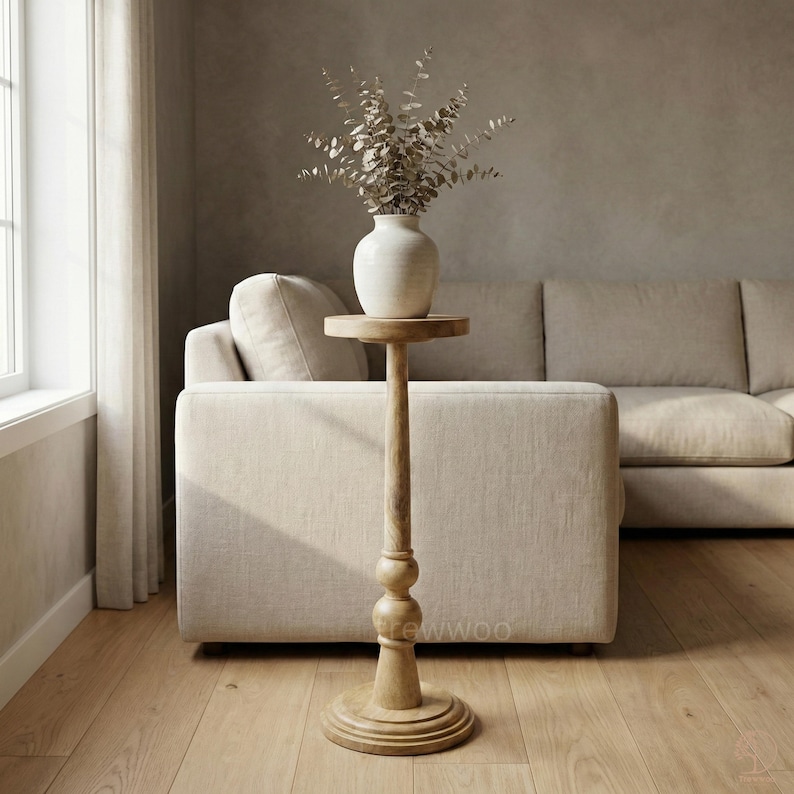 May include: A tall, light-colored wooden side table with a round top and base. A white ceramic vase with dried foliage sits on the table. The table is in front of a beige sofa in a well-lit room.