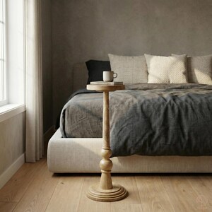 May include: A light brown, wooden side table with a round top and base. The table has a tall, slender stem with a decorative bulbous section. A small white mug and a book rest on the table's surface. The table is next to a bed with a gray comforter.