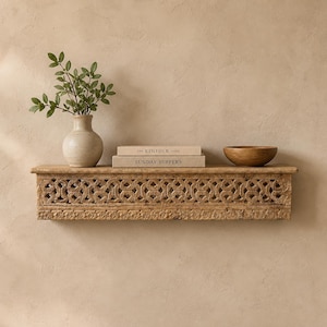 Peut inclure: Étagère en bois sculpté avec un motif géométrique. Elle présente un vase avec des feuilles, deux livres et un bol en bois. L'étagère est fixée à un mur de couleur neutre, créant une ambiance rustique.