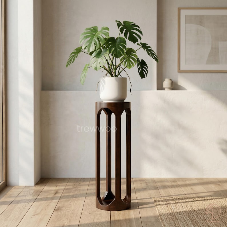 以下が含まれることがあります： 大きな緑の植物が入った白い陶器の鉢が置かれた、濃い茶色の木製プラントスタンド。スタンドは装飾的でオープンなデザインです。「trewwoo」の文字が見えます。プラントスタンドは屋内の木製の床にあります。