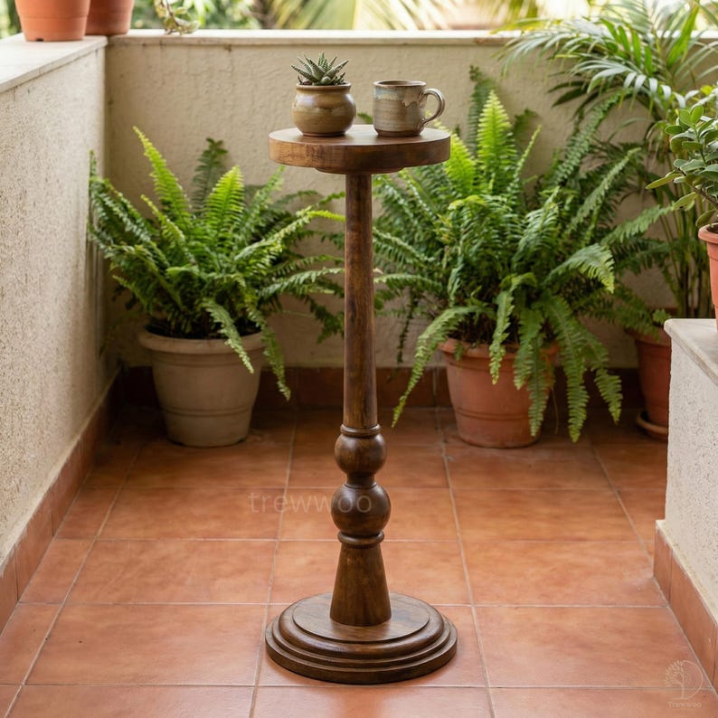 May include: A dark brown wooden plant stand with a round top and base. The stand has a tall, slender stem with decorative bulbous details. A small potted plant and a mug sit on the top. The stand is surrounded by potted ferns.