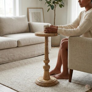 May include: A small, round wooden side table with a pedestal base. The table has a light brown finish and is approximately 24 inches tall. The table is next to a beige armchair and a beige sofa in a living room setting.