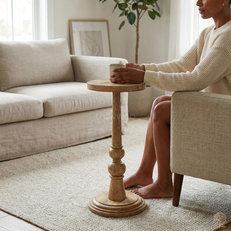 May include: A small, round wooden side table with a pedestal base. The table has a light brown finish and is approximately 24 inches tall. The table is next to a beige armchair and a beige sofa in a living room setting.
