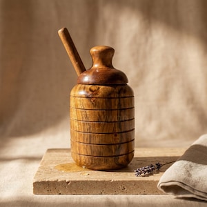 Puede incluir: Un tarro de miel de madera con tapa y cuchara. El tarro cilíndrico tiene ranuras horizontales, y la tapa tiene una parte superior redondeada. La cuchara descansa dentro del tarro. El tarro de miel está sobre una superficie de piedra.