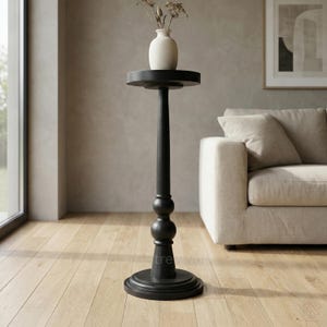 May include: A black wooden plant stand with a circular top and base. A small, cream-colored vase with dried flowers is on the stand. The stand features a decorative, turned design with a bulbous section. The stand is on a wooden floor. The stand is approximately 100 cm tall.