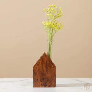 May include: A wooden house-shaped vase with a clear glass tube holding yellow flowers. The vase is made of dark brown wood and has a logo on the front. The flowers have small, round yellow blooms.