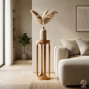 Op de afbeelding: Een lichtgekleurde houten bijzettafel met een vaas met gedroogd pampasgras. De tafel heeft een uniek, open ontwerp met gebogen poten. Bovenop de tafel staat een beige vaas. De kamer heeft een neutraal kleurenpalet met een bank en kunstwerken.