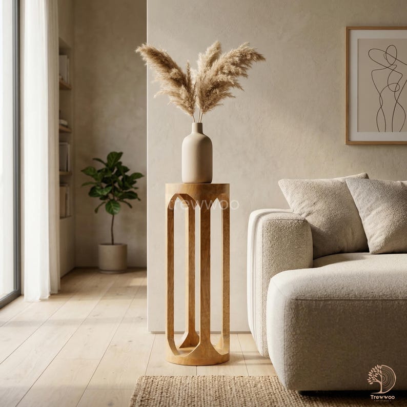 Op de afbeelding: Een lichtgekleurde houten bijzettafel met een vaas met gedroogd pampasgras. De tafel heeft een uniek, open ontwerp met gebogen poten. Bovenop de tafel staat een beige vaas. De kamer heeft een neutraal kleurenpalet met een bank en kunstwerken.