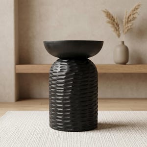 以下が含まれることがあります： ボウル型の天板が付いた、黒色のテクスチャ加工されたアクセントテーブル。 テーブルは、繰り返しのパターンを持つ円筒形のベースを備えています。 テーブルは白いラグの上にあります。