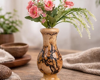 Op de afbeelding: Een decoratieve houten vaas met een uniek, natuurlijk nerfpatroon en een uitlopende bovenkant. De vaas bevat een boeket roze bloemen en groen. De vaas is ongeveer 15 cm hoog en staat op een houten tafel.