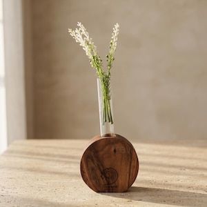 May include: A small, round wooden vase with a clear glass tube holding white flowers. The vase has a dark brown finish and a logo on the front. The flowers have white petals and green stems. The vase is on a light-colored surface.