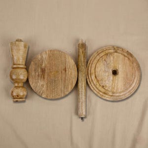 May include: Unassembled wooden table components. The parts include a turned leg, two circular tabletops, and a central support post. The wood has a natural, light brown color and visible grain. The pieces are arranged on a neutral-toned background.