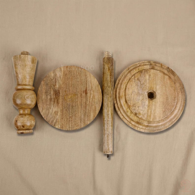 May include: Unassembled wooden table components. The parts include a turned leg, two circular tabletops, and a central support post. The wood has a natural, light brown color and visible grain. The pieces are arranged on a neutral-toned background.