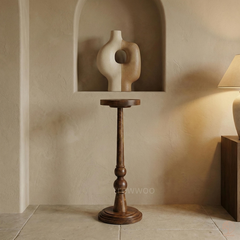 May include: A tall, dark brown wooden plant stand with a round top and base. The stand features a long, slender stem with decorative spherical accents. A two-toned vase with a circular opening sits in a recessed wall niche.