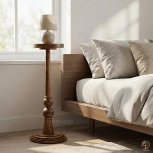 May include: A tall, wooden side table with a small lamp. The table has a round base, a turned column with two decorative spheres, and a small, round tabletop. The lamp has a beige shade and a spherical base. The bed frame and pillows are visible in the background.