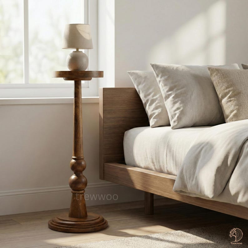 May include: A tall, wooden side table with a small lamp. The table has a round base, a turned column with two decorative spheres, and a small, round tabletop. The lamp has a beige shade and a spherical base. The bed frame and pillows are visible in the background.