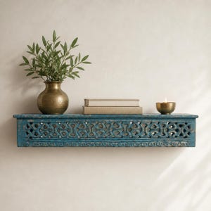 Può includere: Una mensola in legno turchese anticato con dettagli intagliati. Contiene un vaso in ottone con vegetazione, due libri e una piccola ciotola in ottone con una candela accesa. La mensola è montata su una parete colorata neutra.