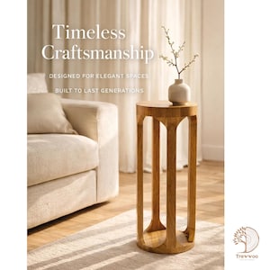 Op de afbeelding: Een lichtbruine houten bijzettafel met een ronde bovenkant en vier verticale steunen. Op de tafel staat een kleine vaas met een takje bloemen. De tekst "Timeless Craftsmanship" is zichtbaar.
