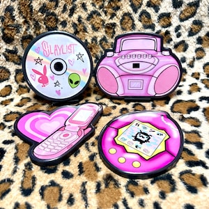 Puede incluir: Cuatro pegatinas rosas con temática de cultura pop sobre un fondo con estampado de leopardo. Las pegatinas incluyen un CD con el texto "SLAYLIST", una radio, un teléfono plegable y un dispositivo estilo Tamagotchi. Cada pegatina tiene un contorno negro.