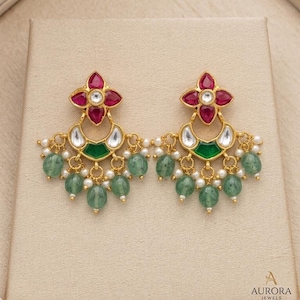 May include: Gold-toned earrings with a floral design featuring red and white accents. The earrings have a green and white crescent shape with dangling green beads and small white pearls. The earrings are displayed on a beige surface.