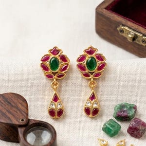 May include: A pair of gold-toned earrings featuring a central oval green stone surrounded by petal-shaped red stones, with a dangling teardrop-shaped charm containing red and white stones. The text Jaipur Crafts is visible at the bottom right.