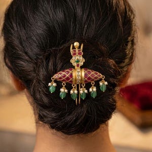 Peut inclure: Un accessoire de coiffure doré doté d'une pièce centrale allongée avec un motif en treillis rouge, agrémenté de petites perles blanches et de perles vertes en forme de goutte d'eau. L'objet inclut une tige verticale avec des incrustations.
