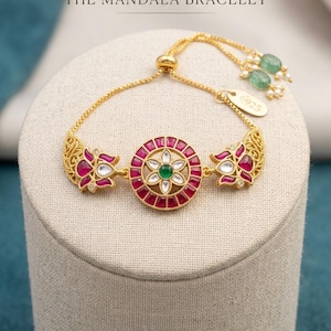 May include: A gold-toned bracelet with a central mandala design featuring a red and white flower pattern with a green center stone. The bracelet has two floral accents and is adorned with green beads and small white pearls. The text "THE MANDALA BRACELET" is above the bracelet.