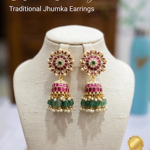 May include: Gold-tone Jhumka earrings featuring a circular top with a central green stone surrounded by red stones and white accents. The bell-shaped base is adorned with red stones, green beads, and small white pearls. Text reads Kundan Heritage.