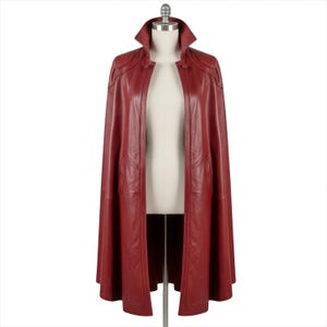 May include: A red leather cape with a high collar and a button closure. The cape is displayed on a mannequin and has a flowing design. The color is a deep, rich red, and the material appears to be smooth and supple.