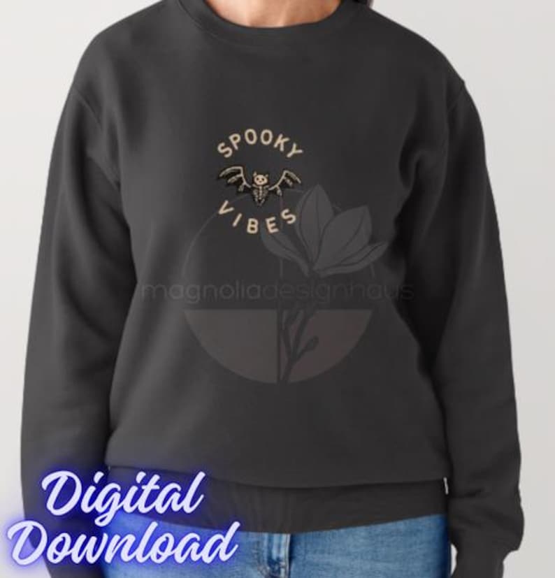 Pu&ograve; includere: Felpa grigio scuro con le parole "Spooky Vibes" e una grafica a pipistrello. Il design include un elemento floreale e il testo "magnoliadesignhaus". Sono visibili anche le parole "Digital Download".