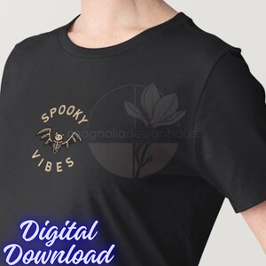 Pu&ograve; includere: T-shirt nero con un design "Spooky Vibes" con una grafica di pipistrello scheletro. Il testo "Digital Download" &egrave; visualizzato in un carattere blu corsivo. Un logo floreale &egrave; visibile sul lato destro della maglietta.