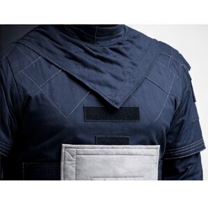 May include: Navy blue quilted jacket with a high collar and diamond stitching. Features black velcro patches and a light gray panel on the lower front. The jacket has a structured design and a modern aesthetic.