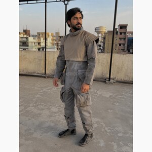 Pu&ograve; includere: Tuta grigia con collo alto e spallina beige. La tuta ha pi&ugrave; tasche e viene indossata con scarpe nere. L'outfit sembra essere un costume o un capo di abbigliamento di una produzione di fantascienza o a tema militare.