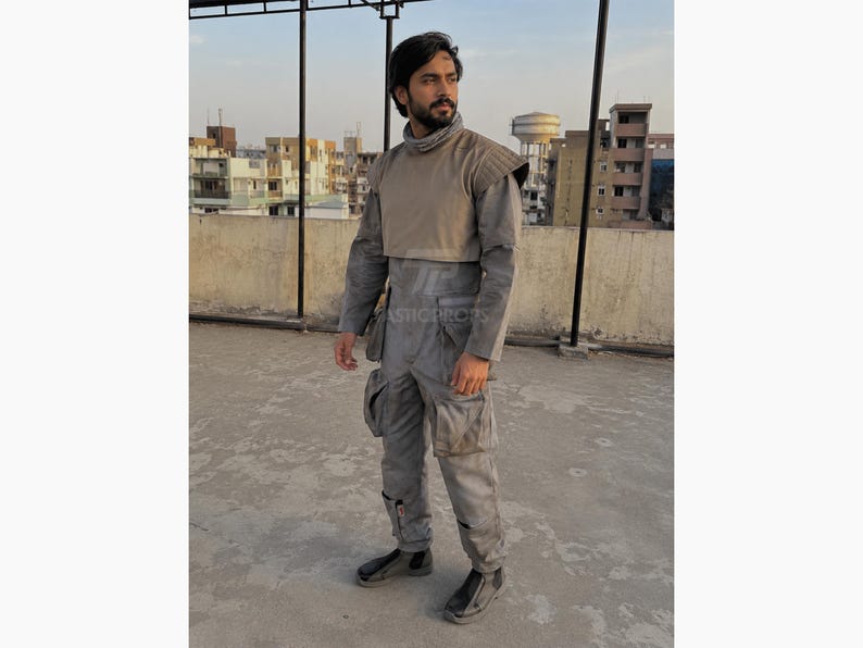 Pu&ograve; includere: Tuta grigia con collo alto e spallina beige. La tuta ha pi&ugrave; tasche e viene indossata con scarpe nere. L'outfit sembra essere un costume o un capo di abbigliamento di una produzione di fantascienza o a tema militare.